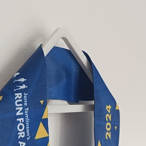 Puede incluir: Una medalla de cinta azul con el texto "Jane Tomlinson's RUN FOR ALL 2024" se muestra en una percha blanca. La cinta tiene acentos de triángulos amarillos y siluetas de personas corriendo.