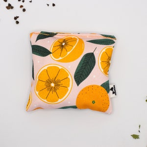 Puede incluir: Una bolsa cuadrada de tela rosa con un estampado repetido de cítricos naranjas, hojas verdes y puntos blancos. La bolsa tiene una pequeña etiqueta en el lateral. Los bordes están ribeteados con tela naranja.