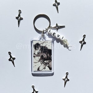 May include: A keychain featuring a black and white image of a character from the anime Attack on Titan, with the name "LEVI" spelled out in white beads. The keychain also includes several silver star-shaped charms.