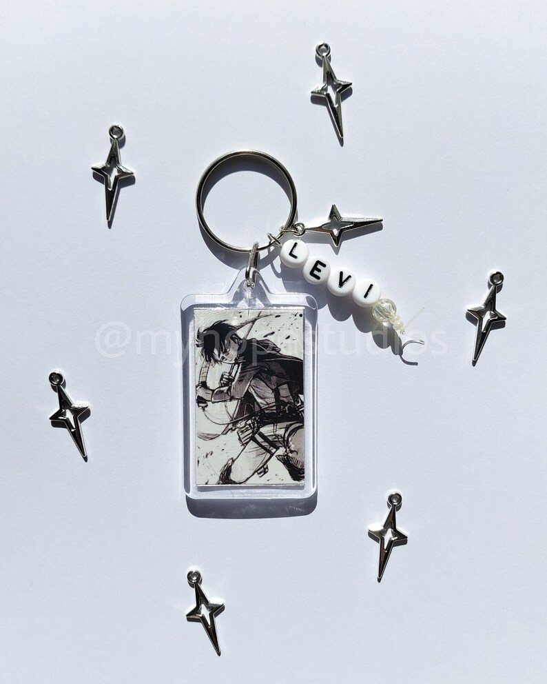 May include: A keychain featuring a black and white image of a character from the anime Attack on Titan, with the name "LEVI" spelled out in white beads. The keychain also includes several silver star-shaped charms.