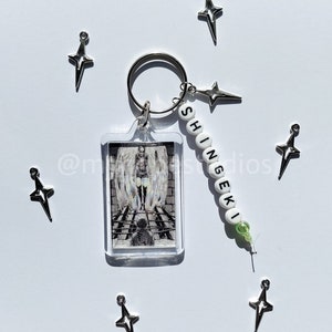 May include: A keychain with a clear acrylic charm featuring a black and white image of a character from the anime "Attack on Titan". The keychain also has a white bead charm with the word "Shingeki" spelled out in black.