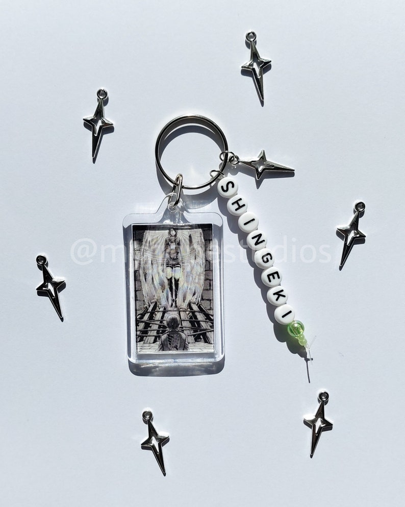 May include: A keychain with a clear acrylic charm featuring a black and white image of a character from the anime "Attack on Titan". The keychain also has a white bead charm with the word "Shingeki" spelled out in black.