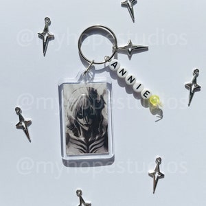 May include: A silver keychain with a clear photo frame featuring a black and white anime character. The keychain also has a silver charm with a star design and a beaded name tag that reads "ANNIE".