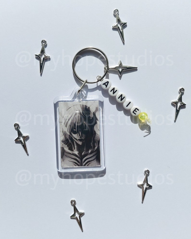 May include: A silver keychain with a clear photo frame featuring a black and white anime character. The keychain also has a silver charm with a star design and a beaded name tag that reads "ANNIE".