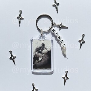 May include: A silver keychain with a clear plastic charm featuring a black and white image of a person. The keychain also has a silver star charm and a white beaded charm with the word "CHANGE" spelled out in black letters.