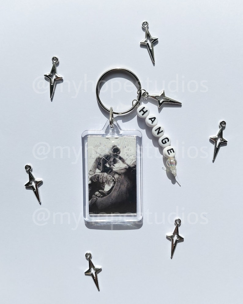 May include: A silver keychain with a clear plastic charm featuring a black and white image of a person. The keychain also has a silver star charm and a white beaded charm with the word "CHANGE" spelled out in black letters.