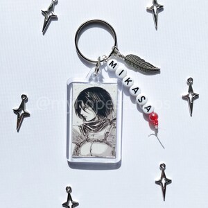 May include: A keychain featuring a clear rectangular photo frame with a black and white image of a character from the anime Attack on Titan. The keychain also includes a silver feather charm, a silver star charm, and white beads with the name "Mikasa" spelled out in black.