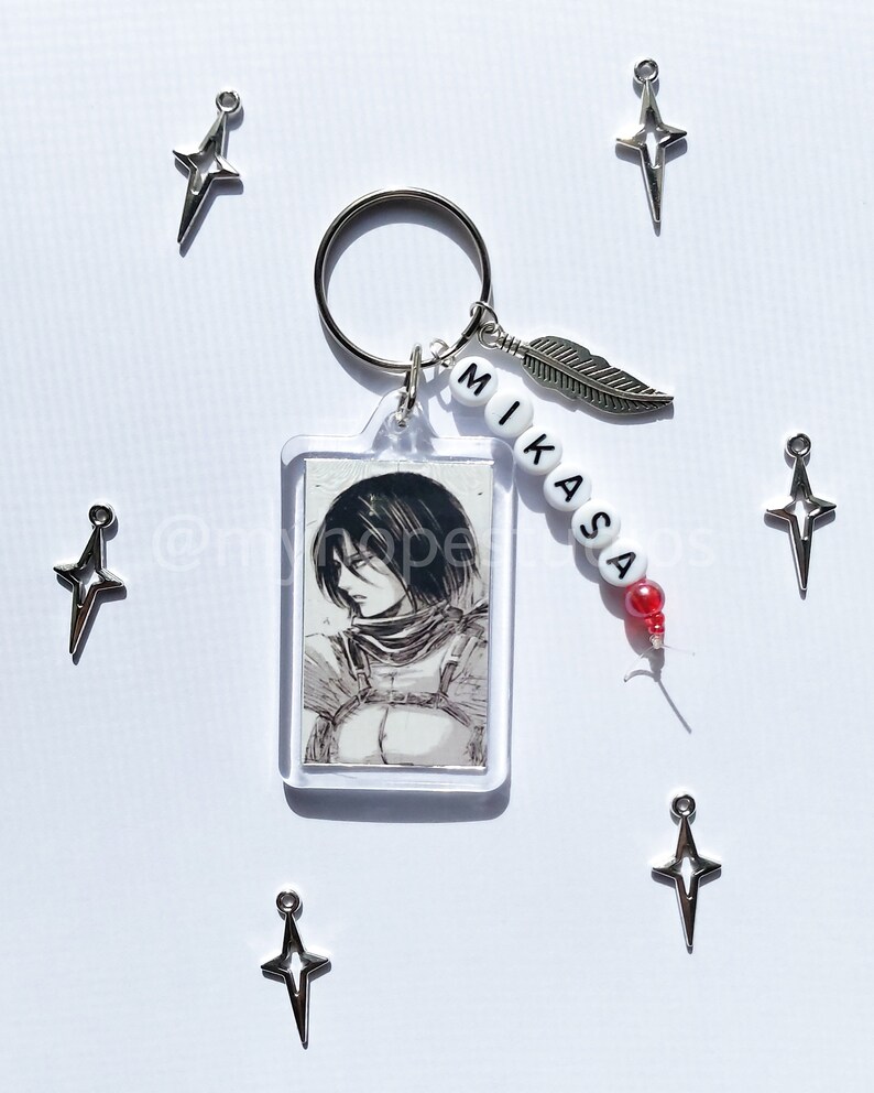 May include: A keychain featuring a clear rectangular photo frame with a black and white image of a character from the anime Attack on Titan. The keychain also includes a silver feather charm, a silver star charm, and white beads with the name "Mikasa" spelled out in black.