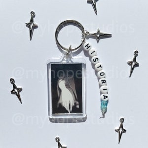May include: A silver keychain with a clear plastic frame holding a black and white photo. The keychain also has a silver star charm and a beaded charm with the word "HISTORIA" spelled out in white.