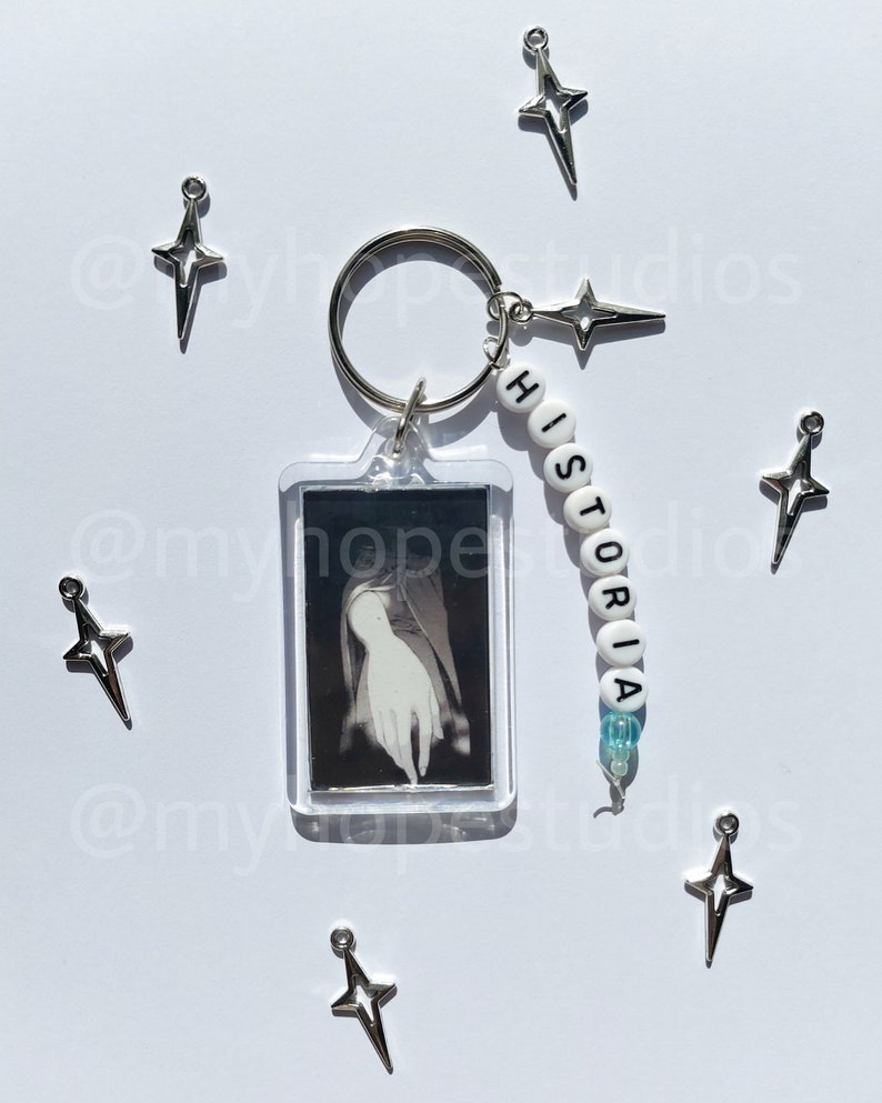 May include: A silver keychain with a clear plastic frame holding a black and white photo. The keychain also has a silver star charm and a beaded charm with the word "HISTORIA" spelled out in white.