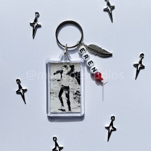 May include: A silver keychain with a clear plastic charm featuring a black and white anime character. The keychain also has a silver feather charm and a beaded name tag that reads "EREN".