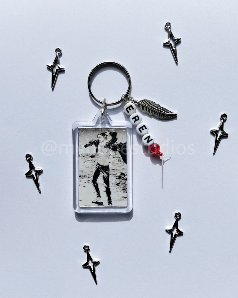 May include: A silver keychain with a clear plastic charm featuring a black and white anime character. The keychain also has a silver feather charm and a beaded name tag that reads "EREN".