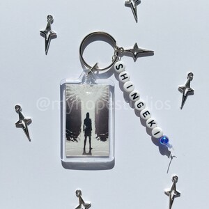 May include: A white keychain with the word "SHINGEKI" spelled out in black beads. The keychain also features a clear plastic charm with a black and white image of a person standing in a dark forest. The keychain is surrounded by silver star-shaped charms.