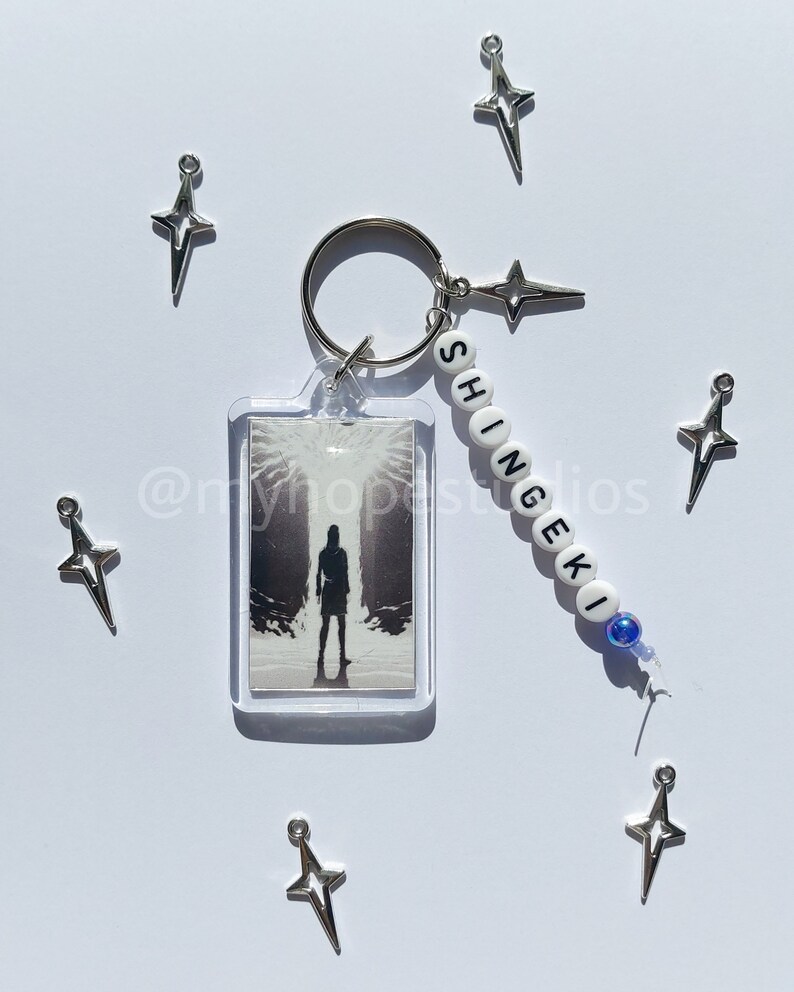 May include: A white keychain with the word "SHINGEKI" spelled out in black beads. The keychain also features a clear plastic charm with a black and white image of a person standing in a dark forest. The keychain is surrounded by silver star-shaped charms.