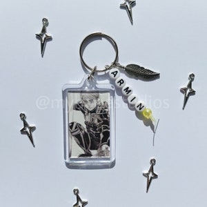 May include: A keychain featuring a character from the anime Attack on Titan. The keychain has a clear plastic frame with a black and white image of the character. The keychain also has a silver charm with a feather and a beaded charm with the word "ARMIN" spelled out in white letters.