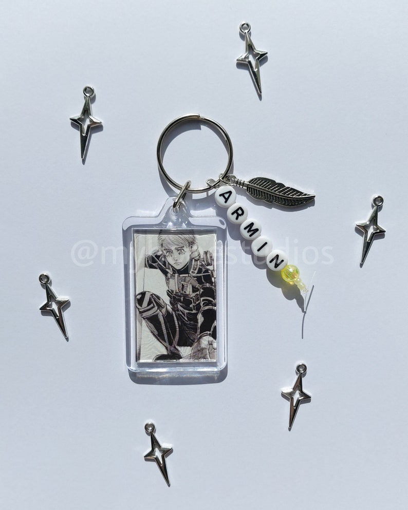 May include: A keychain featuring a character from the anime Attack on Titan. The keychain has a clear plastic frame with a black and white image of the character. The keychain also has a silver charm with a feather and a beaded charm with the word "ARMIN" spelled out in white letters.