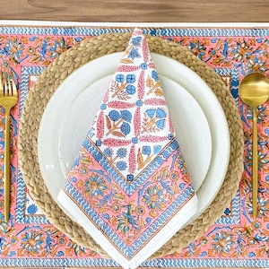 May include: A white placemat with a pink and blue floral pattern, a woven placemat, a white plate, a folded white napkin with a pink and blue floral pattern, a gold fork, and a gold spoon.
