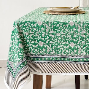 May include: A green tablecloth with a floral pattern and a brown and white striped border. The tablecloth is laid on a table with two chairs and a plant in a white pot.