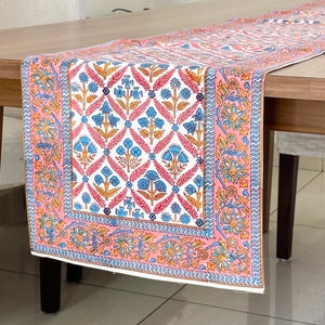 May include: A pink and white table runner with a floral pattern. The runner has a blue and orange border. The runner is on a wooden table.