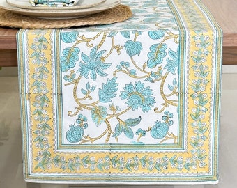 May include: A rectangular table runner with a floral pattern in turquoise and gold on a white background. The runner is displayed on a wooden table, with place settings and decorative items visible. The overall theme is home decor.