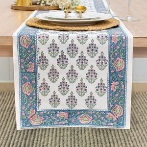 May include: A white table runner with a blue and pink floral pattern. The runner is laid out on a wooden table with two wine glasses, a plate, and silverware.