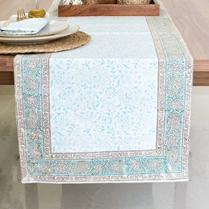 May include: A light blue table runner with a white floral pattern and a brown border. The runner is on a wooden table with two wine glasses, a plate, and a small potted plant.