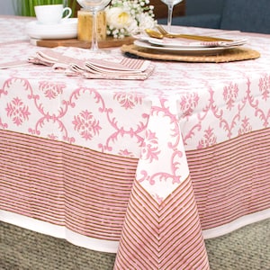 May include: A pink and white tablecloth with a floral pattern and a brown striped border. The tablecloth is laid on a wooden table with two wine glasses and a plate of flowers.
