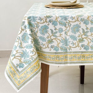 May include: A white tablecloth with a blue and yellow floral pattern. The tablecloth is laid on a wooden table with two wine glasses and a plate on top.