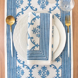 May include: A white table runner with a blue and white floral pattern. The runner has a blue and white striped border. A white plate with a blue napkin is on top of the runner. A gold fork and spoon are on either side of the plate.
