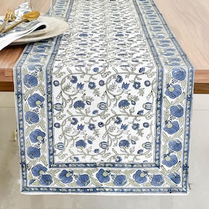 May include: A white and blue floral patterned table runner on a wooden table. The runner features a repeating design of blue flowers and green leaves. Wine glasses, plates, and napkins are set on the table.