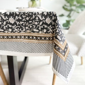 May include: A black and white floral tablecloth with a detailed pattern. The tablecloth features a repeating floral design, a striped border, and a decorative trim. The fabric is draped over a table, showcasing the design and texture of the cloth.