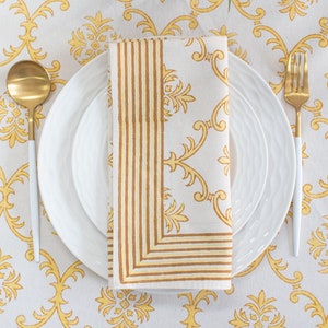 May include: A table setting with a white tablecloth featuring a yellow floral pattern. A folded napkin with a striped border and floral design sits on white plates. Gold-colored utensils and a vase of white flowers complete the elegant arrangement.