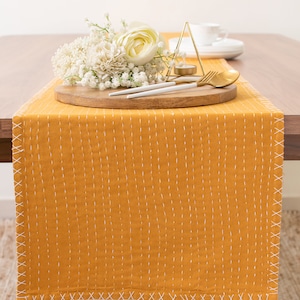 May include: A yellow table runner with white stitching, a wooden tray with white flowers, gold cutlery, and a gold geometric candle holder.