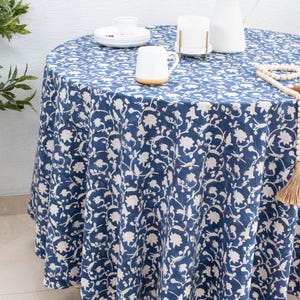 May include: A round tablecloth with a navy blue background and a white floral pattern. The table is set with a white pitcher, a mug, and a plate. A wooden bead garland with a tassel is draped on the table.