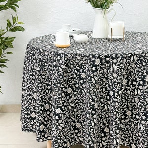 May include: A black and white floral round tablecloth with a white border. The tablecloth is laid on a round table with a white vase of white flowers, a white creamer, a white sugar bowl, and a white teacup and saucer on top.