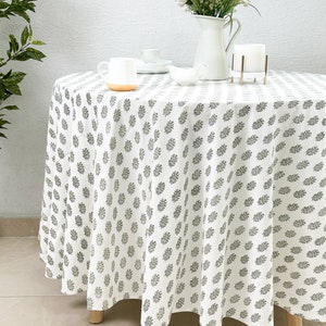 May include: White round tablecloth with a black leaf pattern. The tablecloth is draped over a table with wooden legs. There are white cups and a white jug with white flowers on the table.