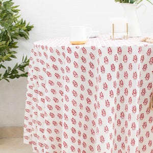 May include: A white round tablecloth with a repeating red floral pattern. A white mug with a gold base sits on the table. A white pitcher with flowers and a gold candle holder are also on the table.