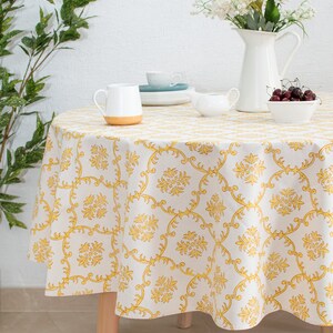 May include: A white round tablecloth with a yellow floral pattern. The tablecloth is on a wooden table with a white pitcher, a white creamer, a white bowl of cherries, and a white saucer with a blue teacup.