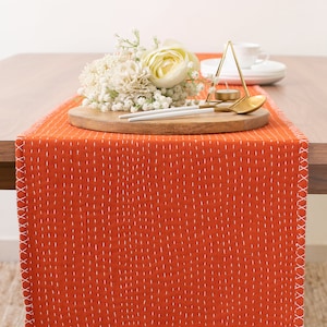 May include: Orange table runner with white stitching, featuring a wooden tray with white flowers and gold utensils.