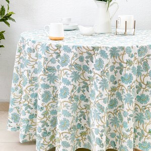 May include: White round tablecloth with a blue and gold floral pattern. The tablecloth is laid on a table with a white pitcher of flowers, two white cups, and a white creamer.
