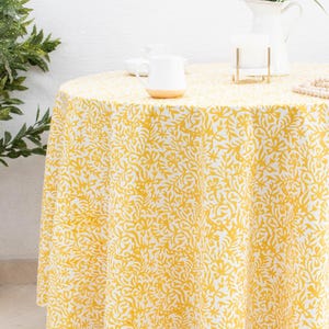 May include: A round tablecloth featuring a yellow and white floral design. The cloth covers a table, with a white mug and a vase of flowers on top. The tablecloth is made from a soft, flowing material, and is suitable for a dining table.