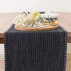 May include: A black quilted table runner with white stitching, featuring a wooden tray with white flowers, a gold fork and spoon, and a gold candle holder on a wooden table.