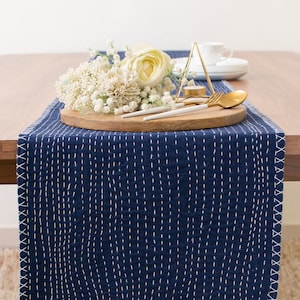 May include: A navy blue table runner with white stitching, a wooden tray with white flowers, a gold fork and spoon, and a gold geometric candle holder on a wooden table.