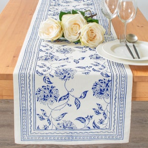May include: A white table runner with a blue floral pattern and a blue border. The runner is decorated with white roses.