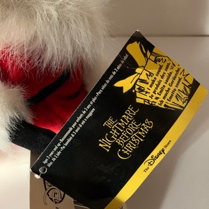 May include: A plush toy from "The Nightmare Before Christmas" with a tag. The toy has white fur, red accents, and black details. The tag is black and yellow with the movie title and "The Disney Store" logo.