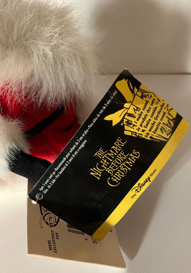 May include: A plush toy from "The Nightmare Before Christmas" with a tag. The toy has white fur, red accents, and black details. The tag is black and yellow with the movie title and "The Disney Store" logo.
