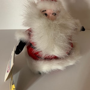 May include: A plush Santa Claus doll with a red hat and suit, trimmed with white faux fur. The doll has a fluffy white beard and a tag attached. The doll is a festive holiday decoration.
