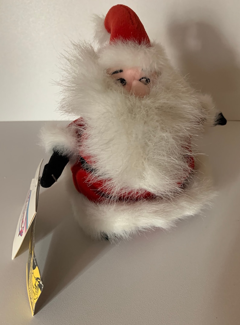 May include: A plush Santa Claus doll with a red hat and suit, trimmed with white faux fur. The doll has a fluffy white beard and a tag attached. The doll is a festive holiday decoration.