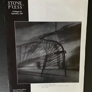 May include: A black and white photograph from the Stone Press Catalogue 24, September 1997. The image features a black wooden chair with a unique, fan-like backrest. The chair is set against a textured wall, with shadows adding depth.