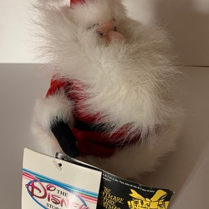 May include: A plush Santa Claus figure with a red hat and suit, white faux fur trim, and black boots. The figure has a tag from The Disney Store, with text that reads "The Nightmare Before Christmas".